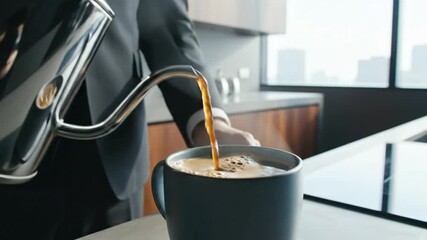 Person in a suit carefully pours coffee from a gooseneck kettle into a mug in a modern kitchen