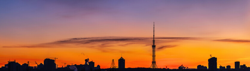 Panoramic view of Kyiv skyline at winter sunset with vibrant orange and purple sky, TV tower silhouette and urban buildings. Cold evening atmosphere, dramatic clouds and city landscape.