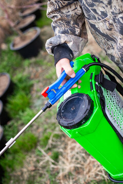A gardener in protective clothing holds a bright green backpack sprayer, preparing to treat young plants in a nursery