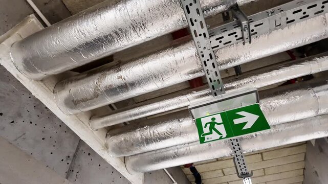 Insulated HVAC pipes and cable tray on mechanical room ceiling with thermal foil wrap, pressure gauge and emergency exit sign &mdash; building services infrastructure, industrial maintenance, no people