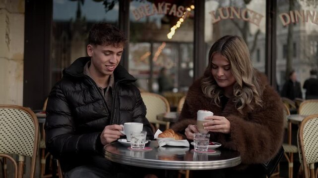 A young couple enjoys coffee and a croissant at an outdoor in Paris France The has a sign that reads Breakfast Lunch Dinner The couple is sitting at a table with chairs enjoying their time together in
