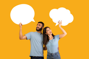 A young arab couple stands together in a studio with a bright yellow background. They hold blank speech bubbles, inviting others to share their thoughts or messages.
