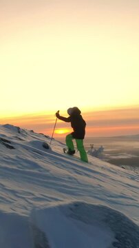 AERIAL Active woman in winter clothes snowshoeing on snowy mountain slope at sunrise. Young woman snowshoe hiking at winter sunset. Traveler snowshoe exploring snowy Lapland mountains in sunny morning
