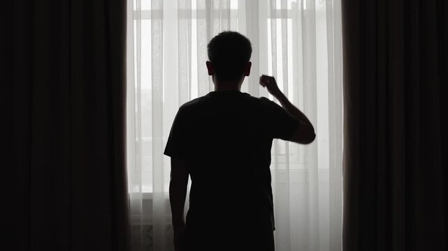 Back view silhouette of stressed man standing by window and holding his head, feeling pressure and uncertainty. Concept of mental overload, difficult decision, anxiety, burnout and life challenges.