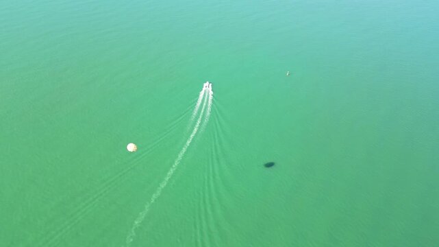 Angled aerial parasailing over green water. 4K horizontal drone footage from above and at slight angle shows a boat in Batumi, Georgia, leaving white wake on emerald sea with a visible parachute.