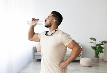 Sporty young Arab man with headphones drinking water after his workout at home. Fit Eastern guy staying hydrated during domestic sports training. Healthy lifestyle, wellness concept