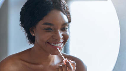 A woman with braces smiles while applying lip gloss to her lips in a bright room. She sits in front of a round mirror. Sunlight comes through the window.