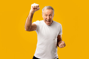 An older man is smiling widely with his fist raised high. He shows excitement and energy in a simple white shirt on a bright yellow background. This scene conveys a sense of joy and victory.