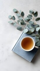 Minimalist Overhead View of a Light Blue Textured Notebook a White Coffee Cup and a Branch of Eucalyptus on a Clean White Desk