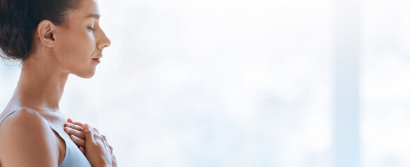 A woman stands in a well-lit room with her eyes closed and hands on her chest. She seems to focus...