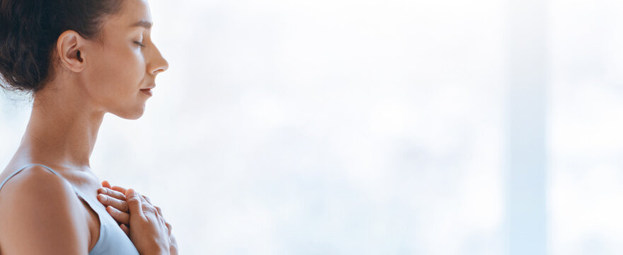 A woman stands in a well-lit room with her eyes closed and hands on her chest. She seems to focus on her breath and inner thoughts. The atmosphere shows calmness and concentration.