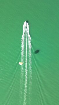 Vertical aerial watersport over green sea. 4K vertical drone footage from top down shows a moving vessel creating white wake, with a parasail flying behind above emerald water in Batumi.
