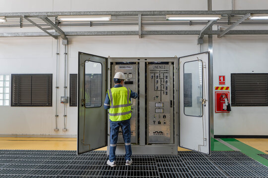 English: Professional electrical engineer standing in front of open switchgear cabinet at power plant, inspecting electrical control system and monitoring energy distribution for building maintenance.
