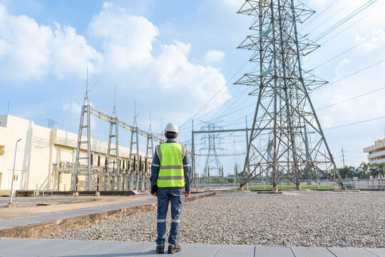 Back view of electrical engineer wearing safety gear looking at high voltage transmission towers and substation, planning power grid maintenance and infrastructure development outdoors.