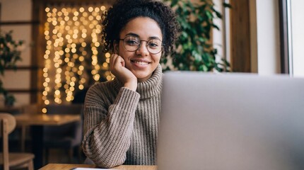 Smiling multiracial woman in glasses and a warm sweater teleworking on a laptop in a trendy cafe with festive bokeh lights background, cozy remote work lifestyle.