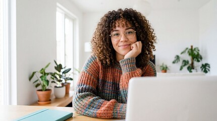 Authentic portrait of a happy young woman with curly hair and glasses sitting at her bright home office desk, working remotely on a laptop with cozy plants around her.