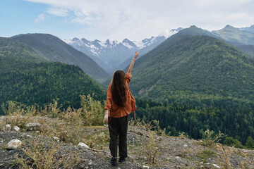 A person stands on a rocky overlook, gazing at vast mountains and green forests, raising an arm toward distant peaks, embracing outdoor adventure.