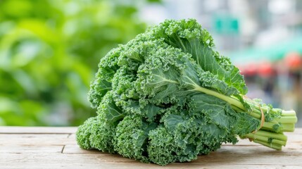 A bundle of fresh kale rests on a wooden table. The vibrant green leaves are crisp and healthy, while a blurred background shows an outdoor market with greenery.