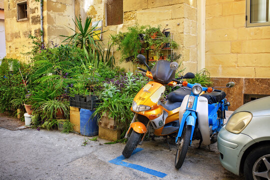 Valletta, Malta - November 05, 2025: Street view with a vintage orange Gilera scooter and a blue Honda parked next to green potted plants by the wall of an old building.
