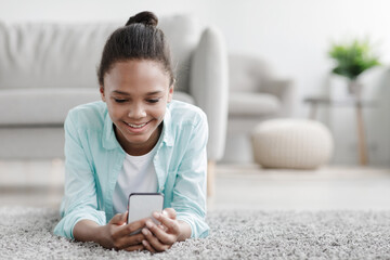 Happy teenage african american girl watch online video, chatting in social networks, lies on floor in living room interior. Texting message in mobile, check app, female relax at home using gadget