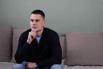 young man sitting on couch at home with hand on chin thinking about solving problems, professional white male with strong facial expression