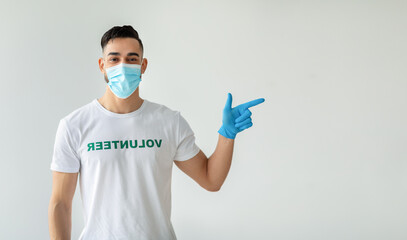 A male volunteer wearing a medical mask stands against a light studio wall, pointing to the side to highlight free space. He promotes an upcoming volunteering event with enthusiasm.