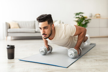 Stay home sports. Handsome young Arab man standing in plank pose with dumbbells, working out indoors. Fit middle Eastern guy strengthening muscles during domestic training in living room