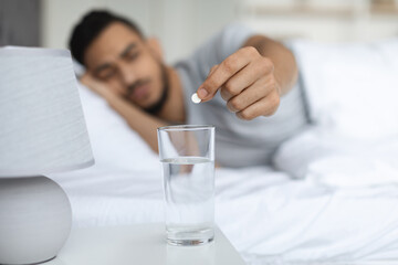 An Arab man prepares to take a pill by placing it into a glass of water on his bedside table. He is sitting on the edge of his bed, indicating discomfort or seeking relief from a headache or insomnia.