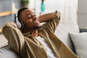 Contented African American Man Listening To Music Wearing Earphones Sitting Holding Hands Behind Head On Sofa At Home. Millennial Guy Relaxing With Eyes Closed Enjoying Playlist. Side View