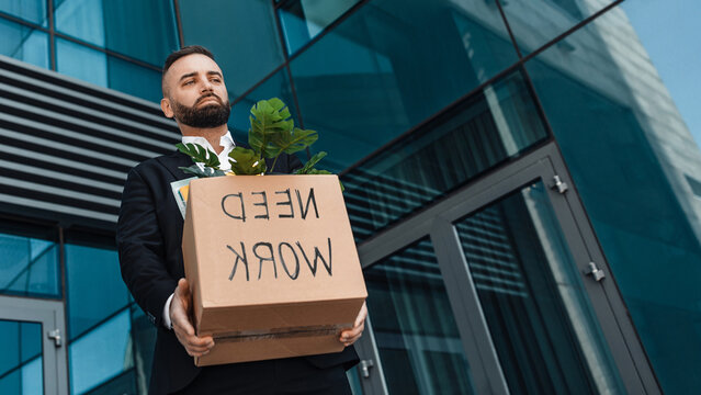 Unemployment concept. Sad businessman with cardboard box of personal stuff and with text of need work, standing near glass office building, panorama with free space