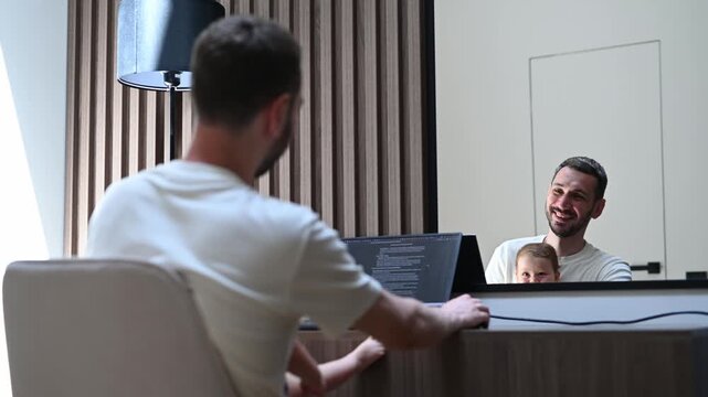 Young father and programmer coding on a laptop while his curious little toddler son secretly approaches and plays with him, showcasing a heartwarming moment of work life balance from home office
