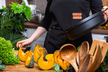 Home cook holding pan with sliced pumpkin in bright cozy kitchen surrounded by fresh green vegetables. Healthy autumn food preparation, cooking routine, lifestyle and blogging concept