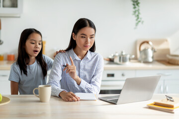 Bored asian girl demanding attention from her busy mother, woman working at home and ignoring her lonely child. Freelancer lady using laptop while sitting in kitchen, copy space