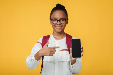 Satisfied smart adolescent black lady pupil in glasses with backpack shows finger at phone with blank screen, isolated on yellow background. App for study, online device, website for remote lesson