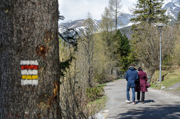 Obraz premium Couple walking on mountain trail with hiking markers on tree. Spring landscape with snow-capped peaks, bare trees, and scenic Alpine path in sunny weather