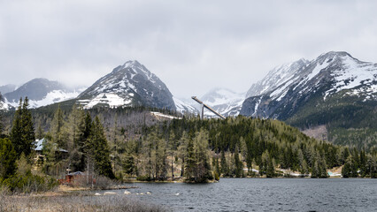 Mountain lake with ski jump tower and snow-capped peaks in High Tatras, Slovakia. Alpine landscape with evergreen forest and dramatic scenery under overcast sky