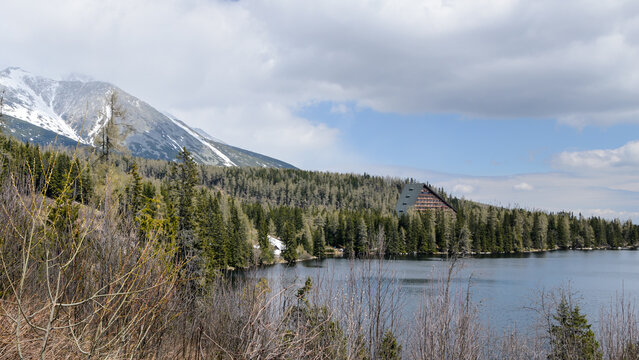 Mountain lake Strbske Pleso in High Tatras, Slovakia. Snow-capped peaks, evergreen forest, historic building on shore. Spring landscape with calm water and dramatic cloudy sky - Powered by Adobe