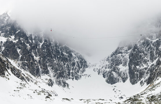 Cable car ascending through misty mountains in High Tatras, Slovakia. Dramatic alpine landscape with snow-covered rocky peaks, glacier valley and fog. Lomnicky stit peak area, winter scenery