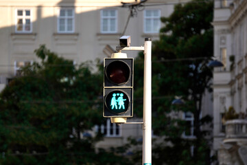 Pedestrian traffic light showing green walk signal, isolated against urban buildings and trees,...
