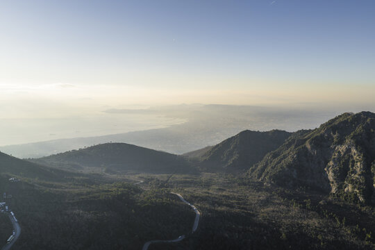 Aerial view of rugged mountain peaks meet the expansive sea under a pale sky, revealing the winding Sentiero del Gran Cono, Ercolano, Campania, Italy.