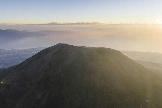 Aerial view of the summit, bathed in the golden light of dawn, overlooking the distant city and the tranquil sea, Vesuvius National Park, Ercolano, Campania, Italy.