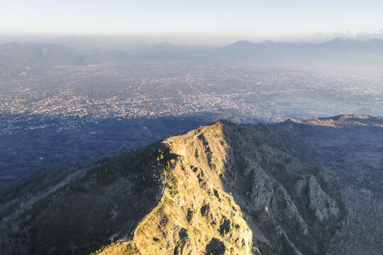 Aerial view of the rugged, sun-kissed peak casting shadows over the vast urban sprawl below, Sentiero del Gran Cono, Ercolano, Campania, Italy.