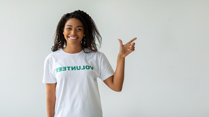 A happy black female volunteer is smiling and pointing to the side, indicating open space to attract attention. She stands against a light wall, embodying a cheerful spirit for volunteering. © Prostock-studio