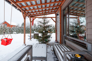 A wooden pergola shelters a snowy porch with a modest Christmas tree, green and white baubles, benches, a table, and a red toddler swing, in a suburban yard.