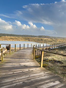 A wooden boardwalk extends over marshy ground beside a tranquil lagoon under a dynamic, cloudy blue sky, near the Furadouro beach and pinhal in Ovar Portugal