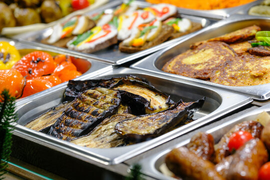 Delicious buffet food spread with grilled eggplant, roasted peppers, and savory pancakes in metal trays.