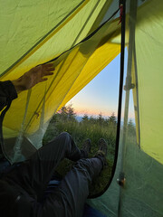 Camper peeks out from yellow tent at dawn, revealing serene view of grassy terrain and distant trees. Dewdrops glisten on tent fabric, with hiking boots placed at tent's entrance.