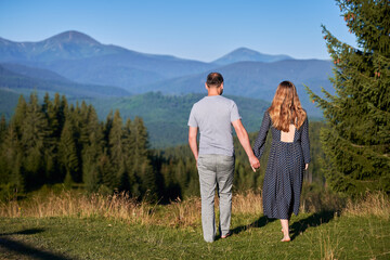 Couple holds hands, walk barefoot through meadow, facing stunning mountain view. Lush green forest and distant mountains under clear blue sky create serene and romantic atmosphere.