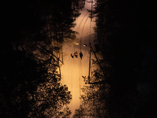 Aerial top view of three people walking along a snowy path through a dark Estonian pine forest illuminated by streetlights