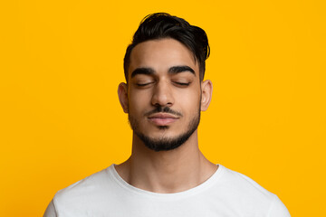 Obraz premium Closeup portrait of handsome bearded millennial middle eastern guy in white t-shirt posing with closed eyes over yellow studio background, enjoying moment, copy space. Male face concept